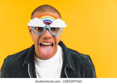 Funny positive young adult man in rainbow mock-up sunglasses sticking his tongue out over yellow background. Studio portrait. High quality photo