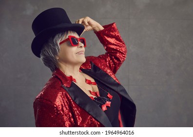 Funny Positive Youthful Looking Grandma Having Fun. Happy Smiling Stylish Retired Senior Woman In Black Tophat, Cool Party Glasses, Jewelry, Bling Red Sequin Concert Outfit Posing On Gray Background