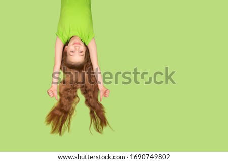 Funny and pretty little girl with extremely long hair smiling isolated on green background. Girl in green T-shirt hanging upside down with flying hair. Copy space.