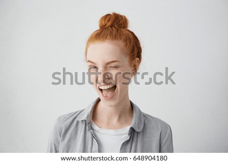 Funny red-haired female teenager with bun wearing casual shirt having joyful expression closing one of her eyes with pleasure and opening widely mouth having broad smile isolated over white background