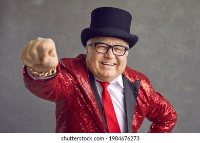 Funny Rich Mature Man In Eyeglasses, Bling Fancy Sequin Jacket And Black Top Hat Smiling And Looking At Camera. Happy Elderly Toastmaster, Magician, Conjurer Or Millionaire Posing On Gray Background