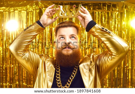 Funny rich young man wearing gold chain and shiny glittering golden jacket putting on king's crown with serious face expression. Ambitious personality, arrogance, megalomania, greed for power concept
