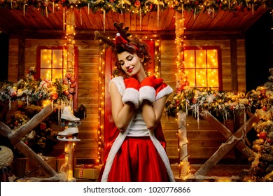 Funny Santa girl in mittens on her hands and deer horns on her head, poses near the house of Santa, decorated with festive lights. Christmas and New Year concept. - Shutterstock ID 1020776866