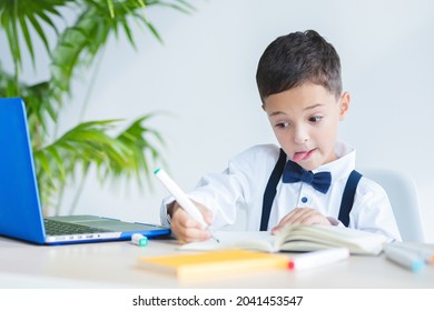 Funny schoolboy, elementary school kid diligently, carefully writes in notebook. Emotional face. Child sits at a table with a computer. Education concept, writing skills, back to school, online lesson