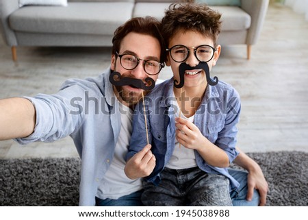 Funny selfie. Happy father and son taking selfie, holding fake moustache on sticks, wearing glasses and smiling at camera, having fun at home, sitting on floor carpet. Parenthood concept
