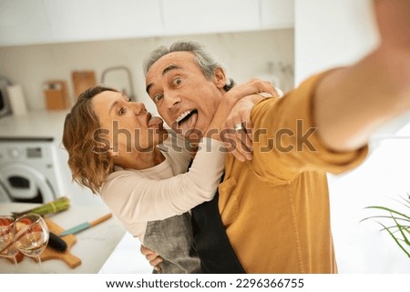 Funny senior couple making selfie, hugging and grimacing, standing in kitchen interior and having fun. Elderly spouses taking photos of happy moments. Retirement lifestyle