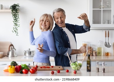 Funny Senior Husband And Wife Having Fun While Cooking At Kitchen, Positive Elderly Man And Woman In Casual Making Healthy Salad And Dancing, Cheerfully Smiling At Camera, Copy Space