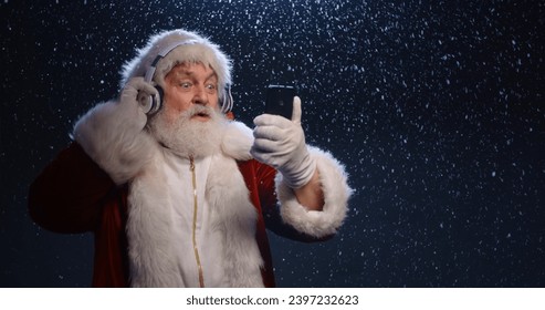 Funny Senior man in santa claus clothes and headphones is dancing to music played on his phone, isolated on blue snowy background - christmas spirit, christmas party close up - Powered by Shutterstock