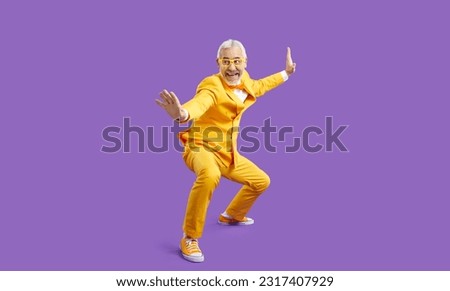 Funny senior man in yellow suit pretends like he knows martial arts. Happy cheerful crazy bearded guy in funky outfit posing in kung fu fighting stance and smiling isolated on solid purple background