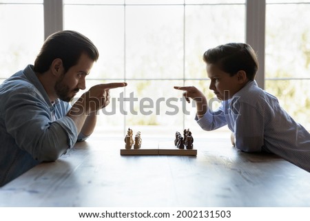 Funny severe dad and little son playing chess, beginning game, sitting opposite like competitors, opponent players at chessboard with start sets, changing comical threatening challenging looks