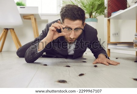 Funny shocked young man lying on white wooden floor in his house, holding his glasses, and looking at lots of disgusting cockroaches crawling everywhere. Concept of roach infestation at home