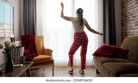 Funny shot of happy woman wearing pajamas and dancing against big window in living room. People relaxing at home, having fun, positive emotions and fun