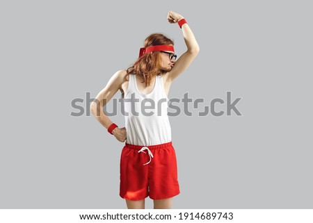 Funny skinny nerd in gym headband, tank top and shorts standing isolated on grey background and kissing weak biceps muscles after his first ever sports workout. Fitness exercise and humour concept