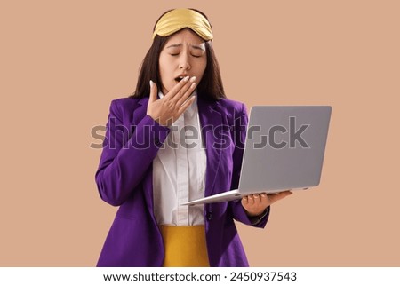 Funny sleepy Asian businesswoman with sleeping mask and laptop on brown background