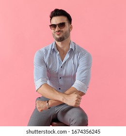 Funny Smart Casual Man Maxing A Duck Face And Holding His Hands In A X Shape, Sitting On A Stool On Pink Studio Background