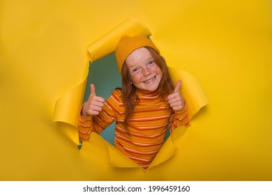 Funny Smiling 7 Year Old Freckled Redhead Girl Breaking Through Yellow Paper Wall With Big Thumbs Up, Ginger Excited Child Says Okay, Like Gesture. Happy Kid Go Back To School. Body Language Concept.