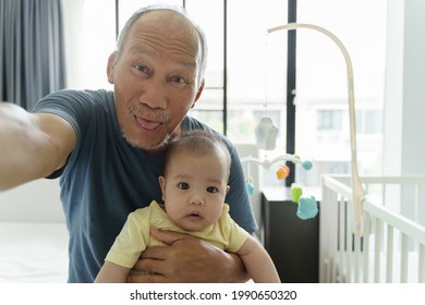 Funny Smiling Asian Grandfather And Little Cute Baby Boy Doing Online Video Call With Family On Mobile Phone. Happy Senior Man And Grandson Taking Selfie Photos With Smartphone At Home.