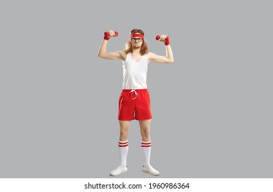 Funny Smiling Hipster Skinny Young Guy In Red And White Sportswear Standing And Doing Workout With Dumbbels Feeling Tired Over Light Grey Wall Background. Funny Active Sport Lifestyle Of Man Concept