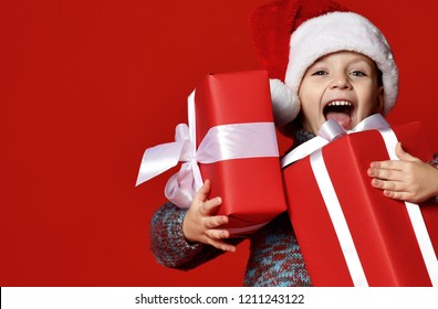 Funny Smiling Joyful  Child Boy In Santa Red Hat Holding Christmas Gift In Hand Over The Red Background