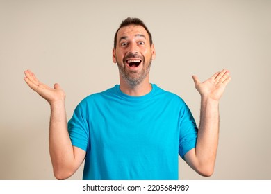 Funny Smiling Man In Blue T-shirt Showing Palms Up, Unexpected Surprise Gesture Isolated On Beige Background. Attractive Happy Man Hearing Good News, Catching Something