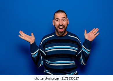 Funny smiling man in striped sweater showing palms, unexpected surprise gesture isolated on blue background. Attractive happy man hearing good news, catching something.