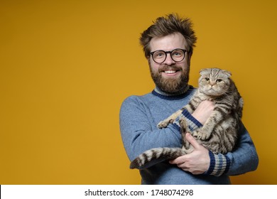 Funny Smiling Shaggy Man Hugs His Adorable Cat, Who Is Not Happy That The Owner Is Holding It In Hands. Copy Space.