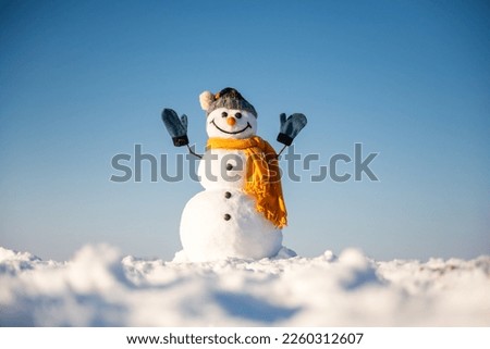 Funny snowman in knitted hat and yellow scalf with hands up on snowy field. Blue sky on background