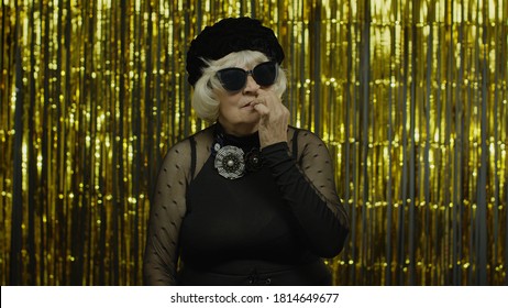 Funny Stupid Senior Old Woman Picking Nose With Silly Brainless Humorous Expression, Removing Boogers, Uncultured Habit, Bad Manners. Elderly Stylish Lady Grandma On Golden Background