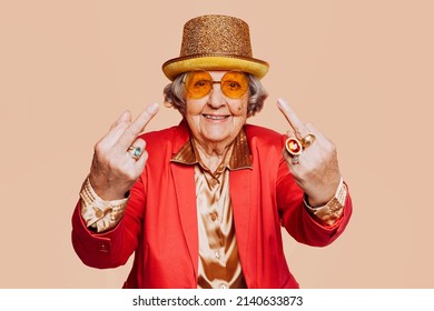 Funny Stylish Elderly Grandmother Portrait Showing Middle Finger Gesture With Hand At Studio. Senior Old Woman Dressing Elegant For A Special Event. Rebel Granny Over Beige Background.