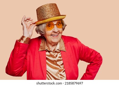 Funny stylish elderly grandmother poses with golden hat and pink suit at studio. Senior old woman looking at cameraf over beige background.