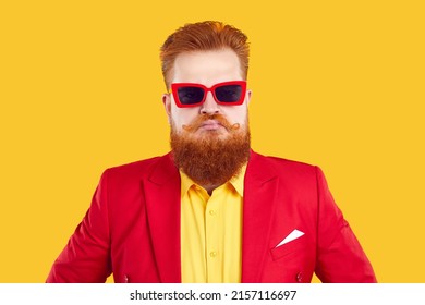 Funny stylish extravagant bearded chubby man pretending to be serious on vivid yellow background. Humorous red-haired guy in red jacket and sunglasses with funny serious and menacing look looks at you