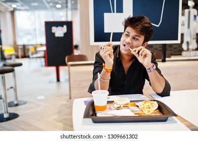 Funny And Stylish Indian Man Sitting At Fast Food Cafe And Eating French Fries, Show His Tongue.