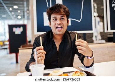 Funny And Stylish Indian Man Sitting At Fast Food Cafe And Eating French Fries, Show His Tongue.