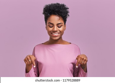 Funny surprised African American female looks with joyful eyes down and indicates as shows something, sees comic things, isolated over lavender wall. Positive amazed young woman poses in studio.