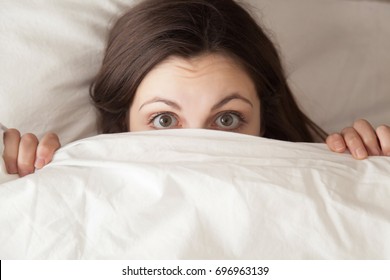 Funny surprised girl covering half of face with white blanket, young scared woman hiding and peeking from duvet, afraid of night monsters, feels embarrassed, wide awake, head shot close up, top view - Shutterstock ID 696963139