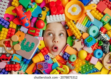 Funny surprised little girl lying in the chaos of toys. Kid's face surrounded by building blocks.
