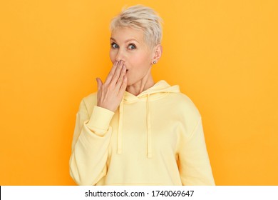 Funny Surprised Mature Woman In Hoodie Covering Mouth With Hand, Having Amazed Astonished Facial Expression, Confused With Intriguing Amazing News. Cute Retired Lady Expressing Astonishment