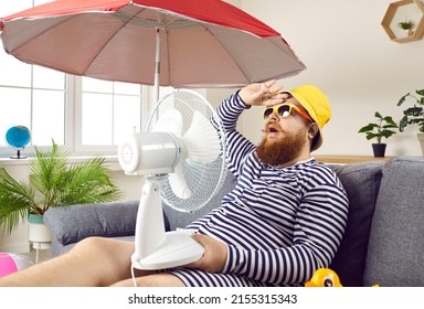 Funny sweaty chubby bearded man in swimsuit sitting at home, suffering from crazy summer heat, wiping sweat off forehead, holding electric fan, wishing for heatwave to stop and fresh breeze to blow - Shutterstock ID 2155315343
