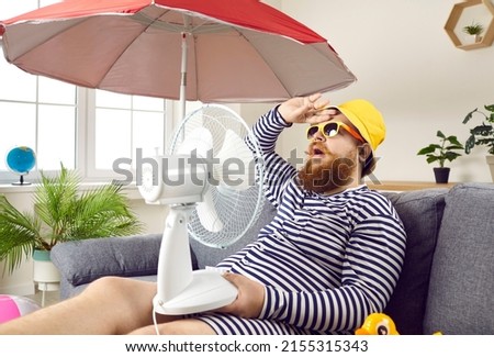 Funny sweaty chubby bearded man in swimsuit sitting at home, suffering from crazy summer heat, wiping sweat off forehead, holding electric fan, wishing for heatwave to stop and fresh breeze to blow