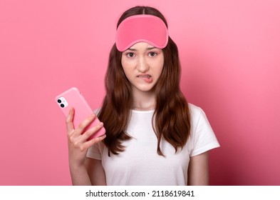 Funny Teen Girl Bit Her Lip Feel Guilty For Oversleep The Alarm Clock Late For School Online Lessons Hold Mobile Phone In Hand Just Wake Up Wearing A Sleep Mask And A White T-shirt On Pink Background