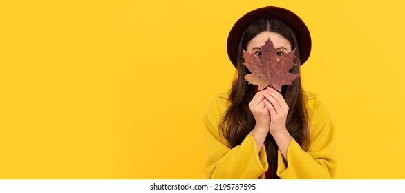 Funny Teen Girl In Hat And Coat Hiding Behind Autumn Maple Leaf On Yellow Background, Season. Autumn Fall Child For Poster Design. Banner Header, Copy Space.