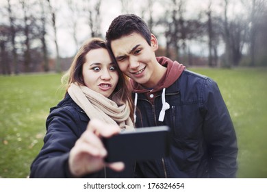 Funny Teenage Couple Photographing Themselves With Smart Phone In The Park. Cute Young Girl With Her Boyfriend Taking Self Portrait With Cell Home Outdoors.