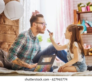 Funny time! Father and his child are playing at home. Cute girl is doing makeup to her dad, sitting on the bed in the bedroom. Family holiday and togetherness.
