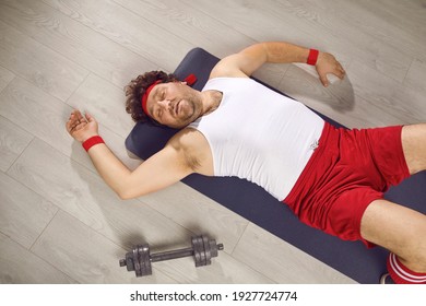 Funny Tired Fat Man Lying On Fitness Mat After Gym Workout. Exhausted Guy In Retro Sweatband, Tank Top And Shorts Fallen On Floor After Sports Exercise With Dumbbells. Top View, High Angle, From Above