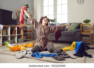 Funny Tired Housewife Crying Desperately While Cleaning Up Terrible Mess At Home. Exhausted Young Woman In Home Clothes Tidying Up Her House And Picking Up Scattered Laundry From Floor In Living-room