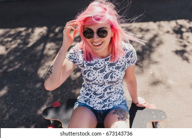 Funny Top View Of Sporty Beautiful Smiling Woman With Blowing Pink Hair Wearing Black Sunglasses, With Tattoo, Sitting On Her Long Board Waiting Her Friends In The Park. Lifestyle And Sport Concept.