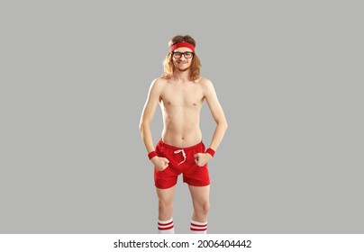 Funny topless skinny man in glasses, red retro sweatband and shorts standing isolated on gray background, grinning, winking eye and showing his weak thin body after first ever sports exercise workout