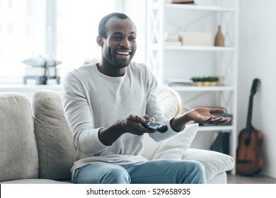 Funny TV Show. Handsome Young African Man Watching TV And Smiling While Sitting On The Sofa At Home 