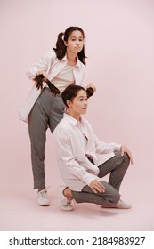 Funny Twin Girls. One Is Playing With Pigtails Of The Other. They Are Wearing White Shirts, Black And White Patterned Pants Over Pink Background.