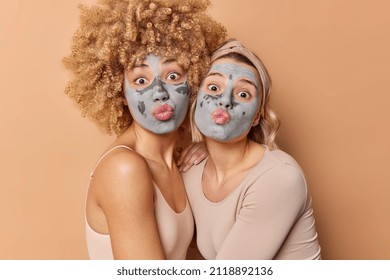 Funny two female models keep lips rounded make funny grimace apply nourishing clay masks on face for rejuvenation and pampering dressed casually isolated over brown background. Beauty concept